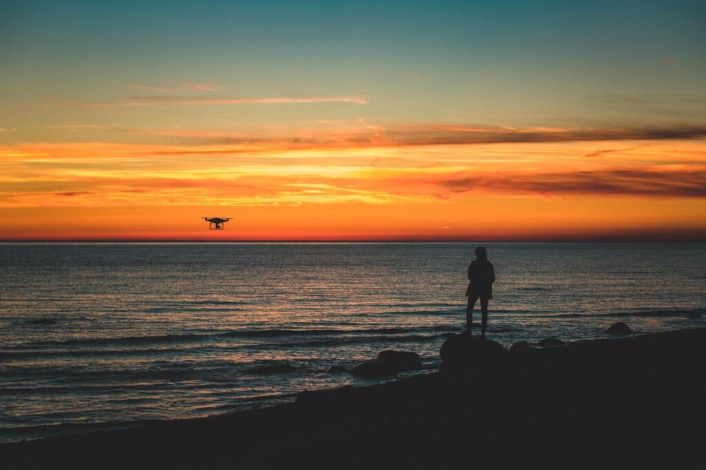 Pilota sulla riva del mare che osserva il drone al tramonto